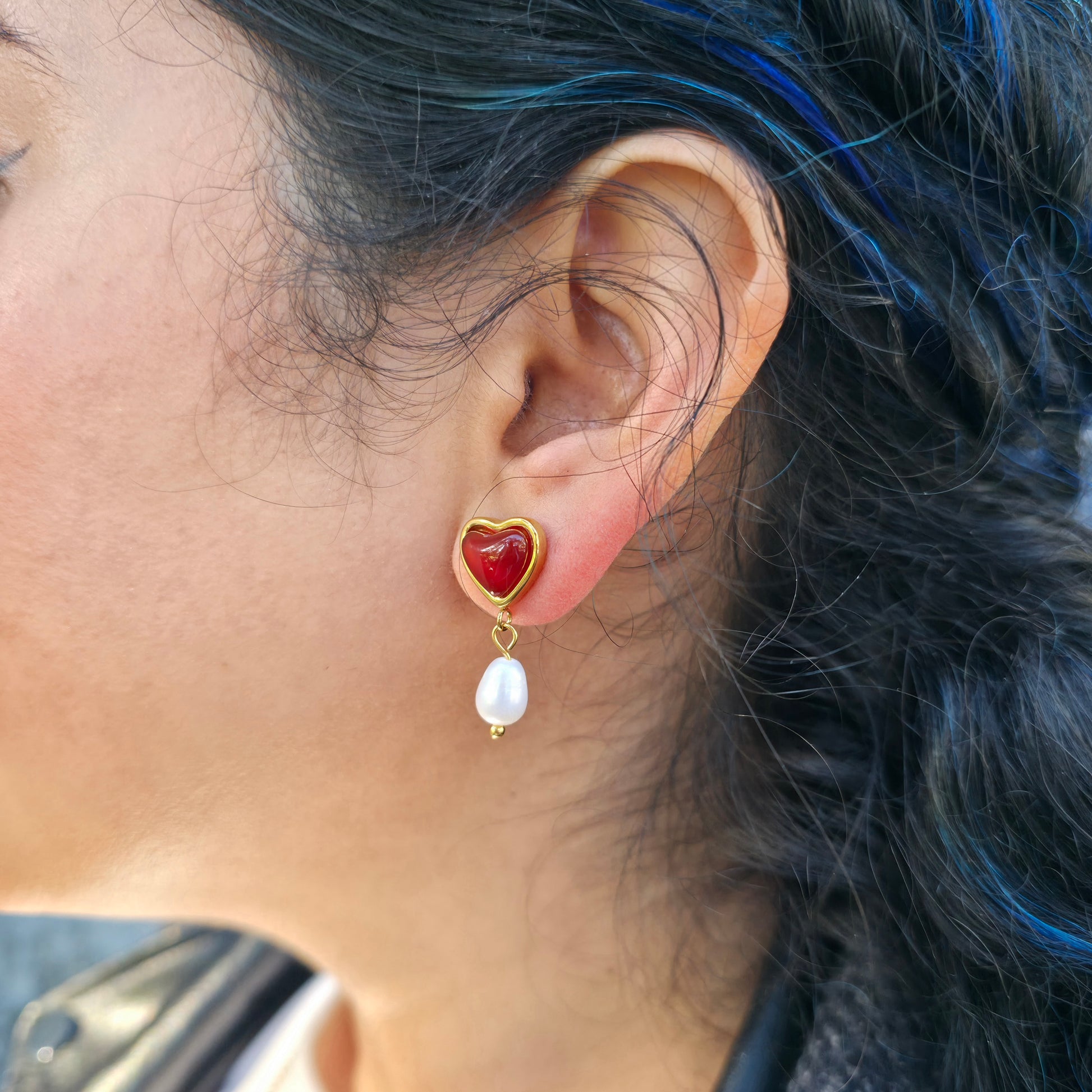 Heart-shaped earring with red stone and pearl drop worn by a person with blue hair.